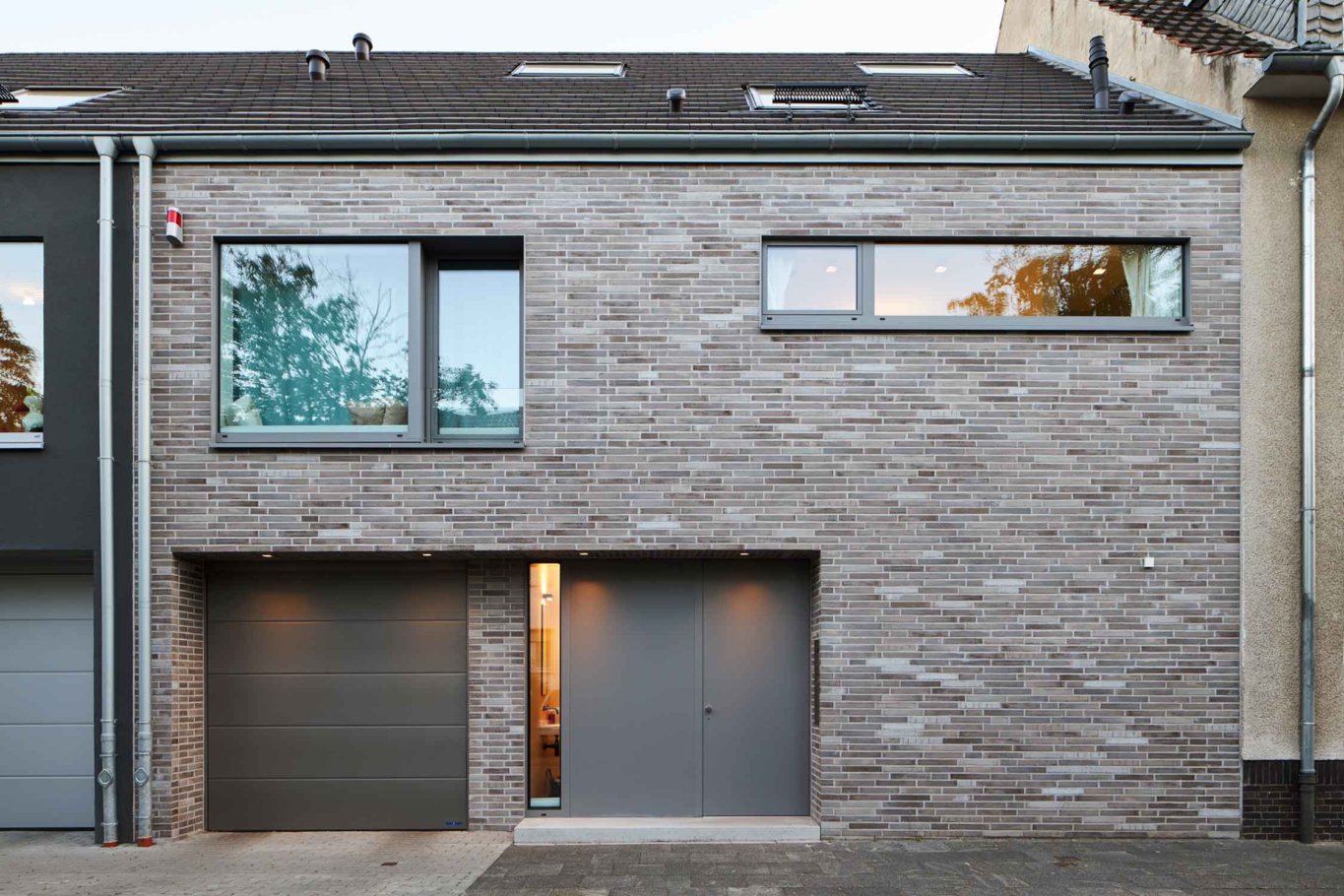 Architekturfotografie Lioba Schneider: Ein Doppeleinfamilienhaus in Bonn bei Dämmerung von der Strasse aus gesehen. Die Fassaden sind unterschiedlich gestaltet- anthrazitfarbener putz und einmal heller Klinker.
