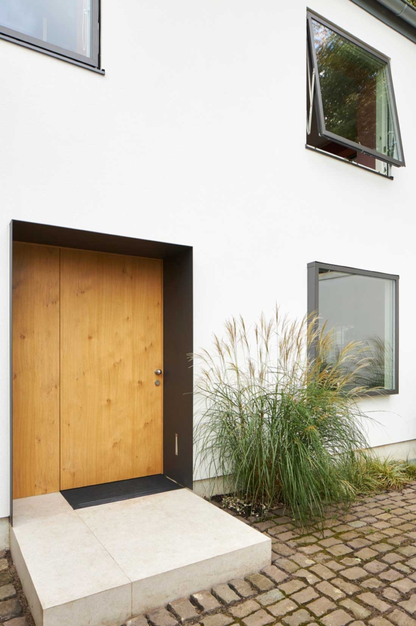Architekturfotografie Lioba Schneider: Umbau eines 60er Jahre-Hauses in Köln. Bündige Fenster in weisser Fassade geben einen modernen Eindruck. Das helle Holz der zurückspringenden Eingangstür sieht hochwertig aus.