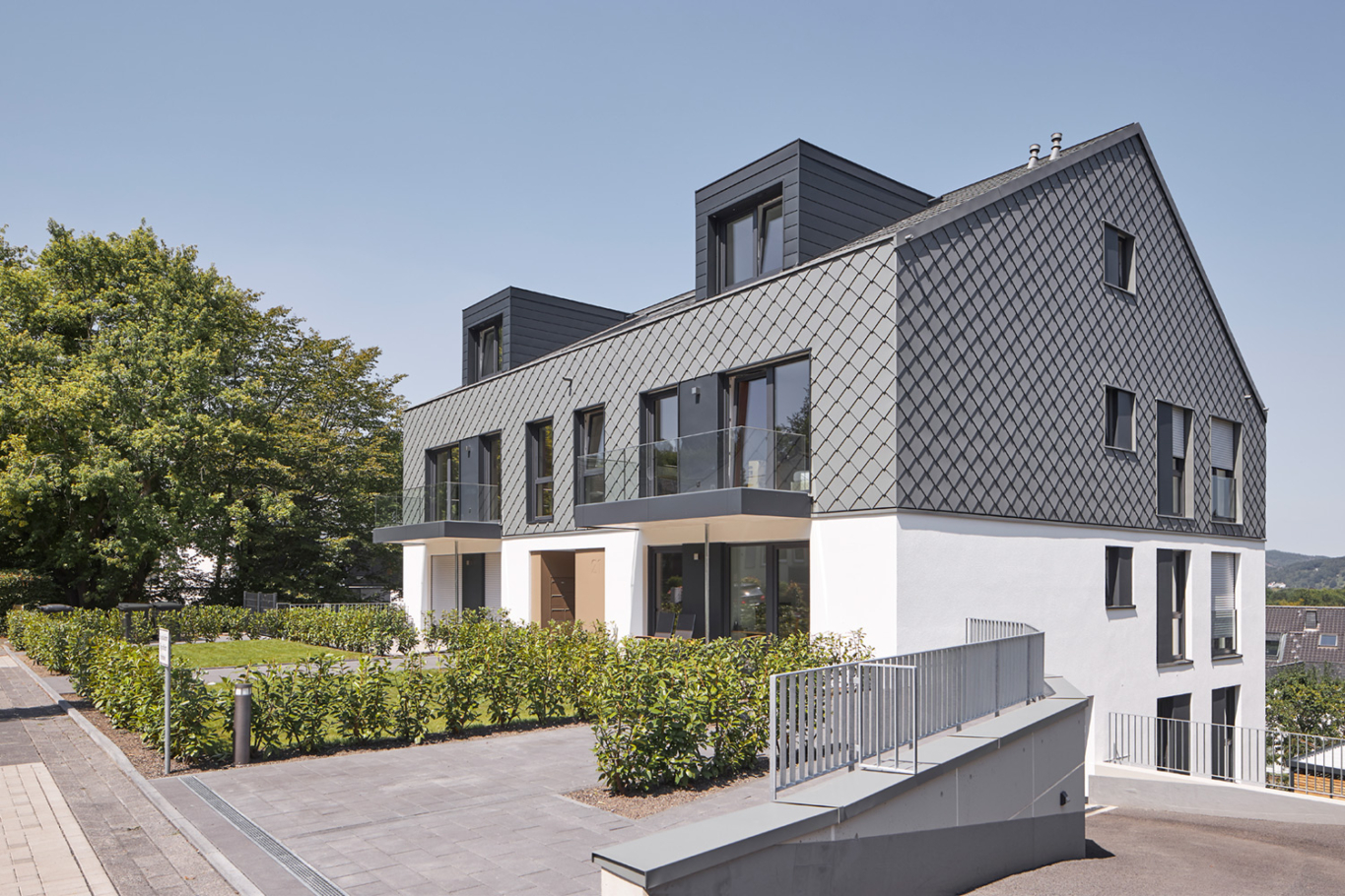 Architekturfotografie: Neubau eines Mehrfamilienhauses mit neun Wohnungen am Hang gebaut. Die Fassade ist teilweise mit grauen Schindeln in Rautenform gestaltet.