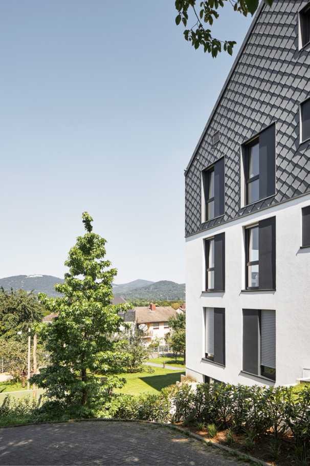 Architekturfotografie: Neubau eines Mehrfamilienhauses mit neun Wohnungen am Hang gebaut. Die Fassade ist teilweise mit grauen Schindeln in Rautenform gestaltet.