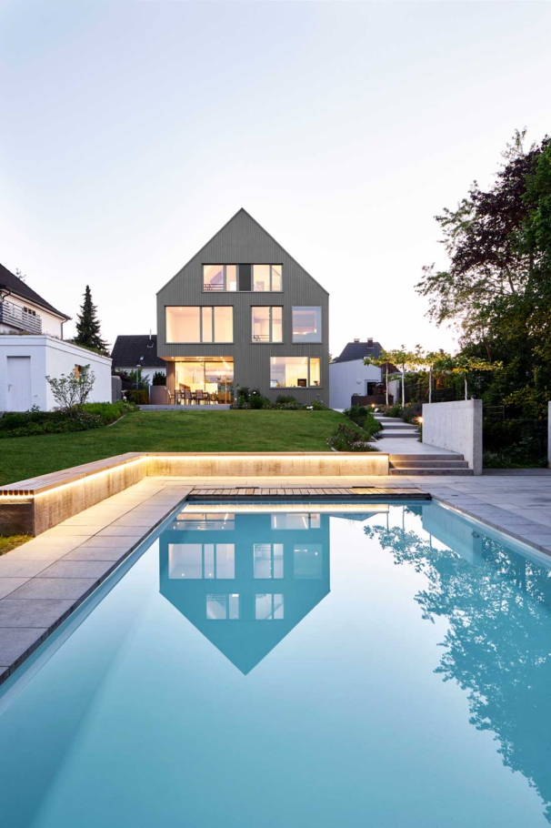 Architekturfotografin Lioba Schneider fotografiert im Auftrag von tr-architekten: Spitzgiebeliges graues Einfamilienhaus in Holzbauweise. Große Fenster lassen viel Licht ins Innere. Im Pool im Garten spiegelt sich das Haus in der Dämmerung.