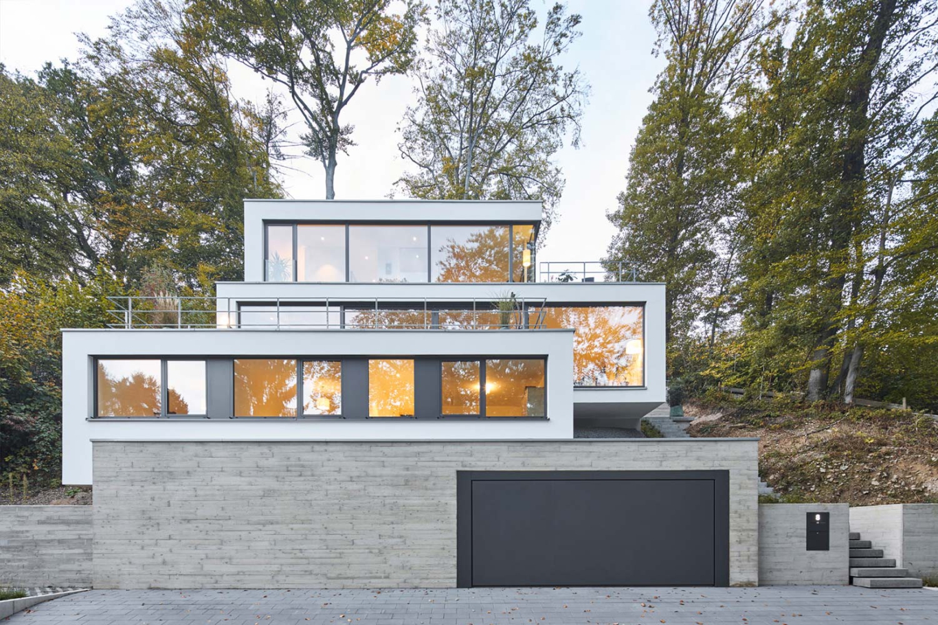 Architekturfotografie Lioba Schneider: Ein weisses Einfamilienhaus mit Flachdach und in mehreren Terrassen schmiegt sich an den Hang im Wald.