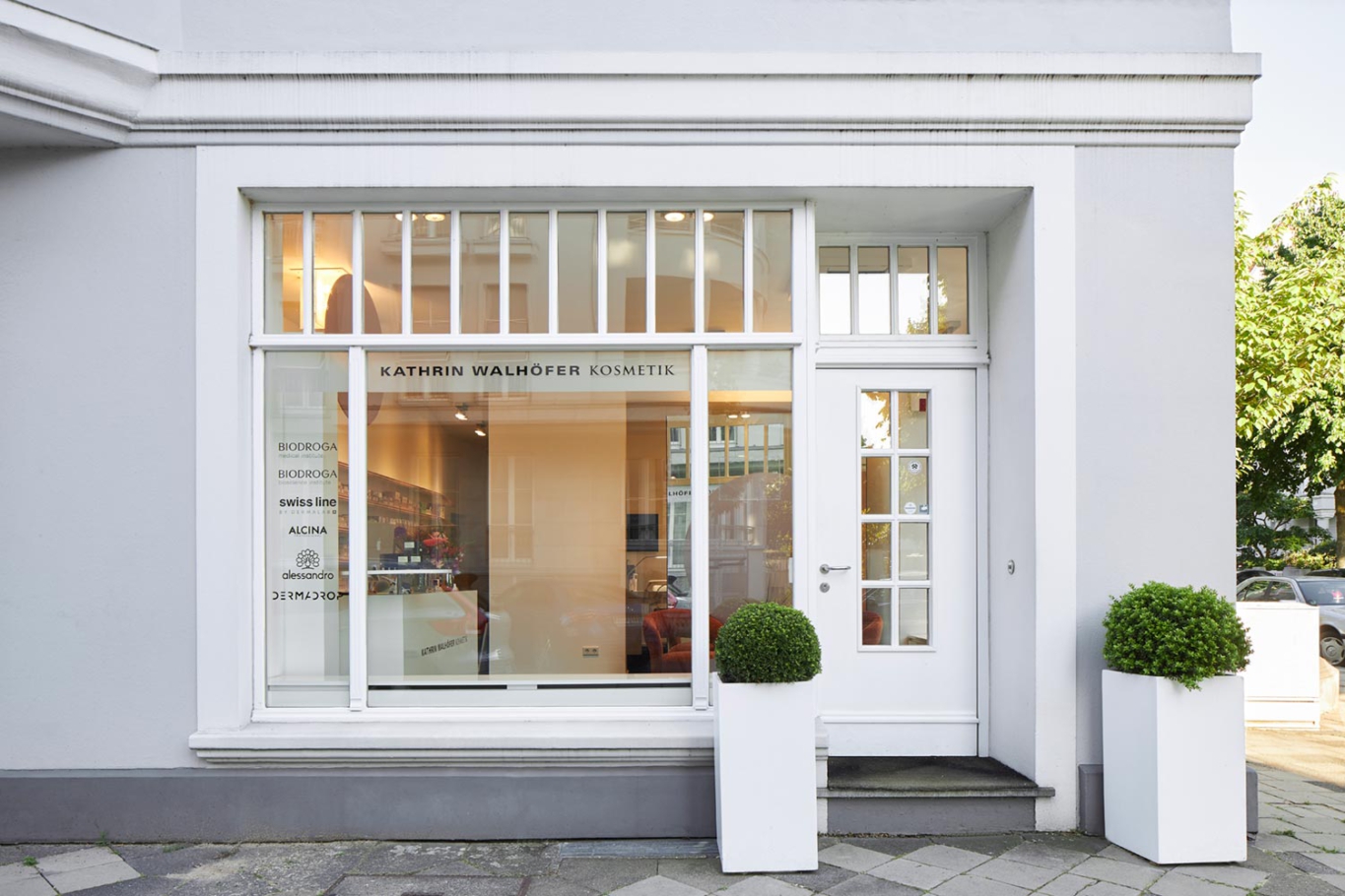 Interiorfotografie Lioba Schneider: Ästhetischer Umbau eines kleinen Kosmetikstudios in Düsseldorf. Die weisse Aussenfassade eines Altbaus zeigt durch ein grosses Fenster den Blick und erleuchtete Innere.