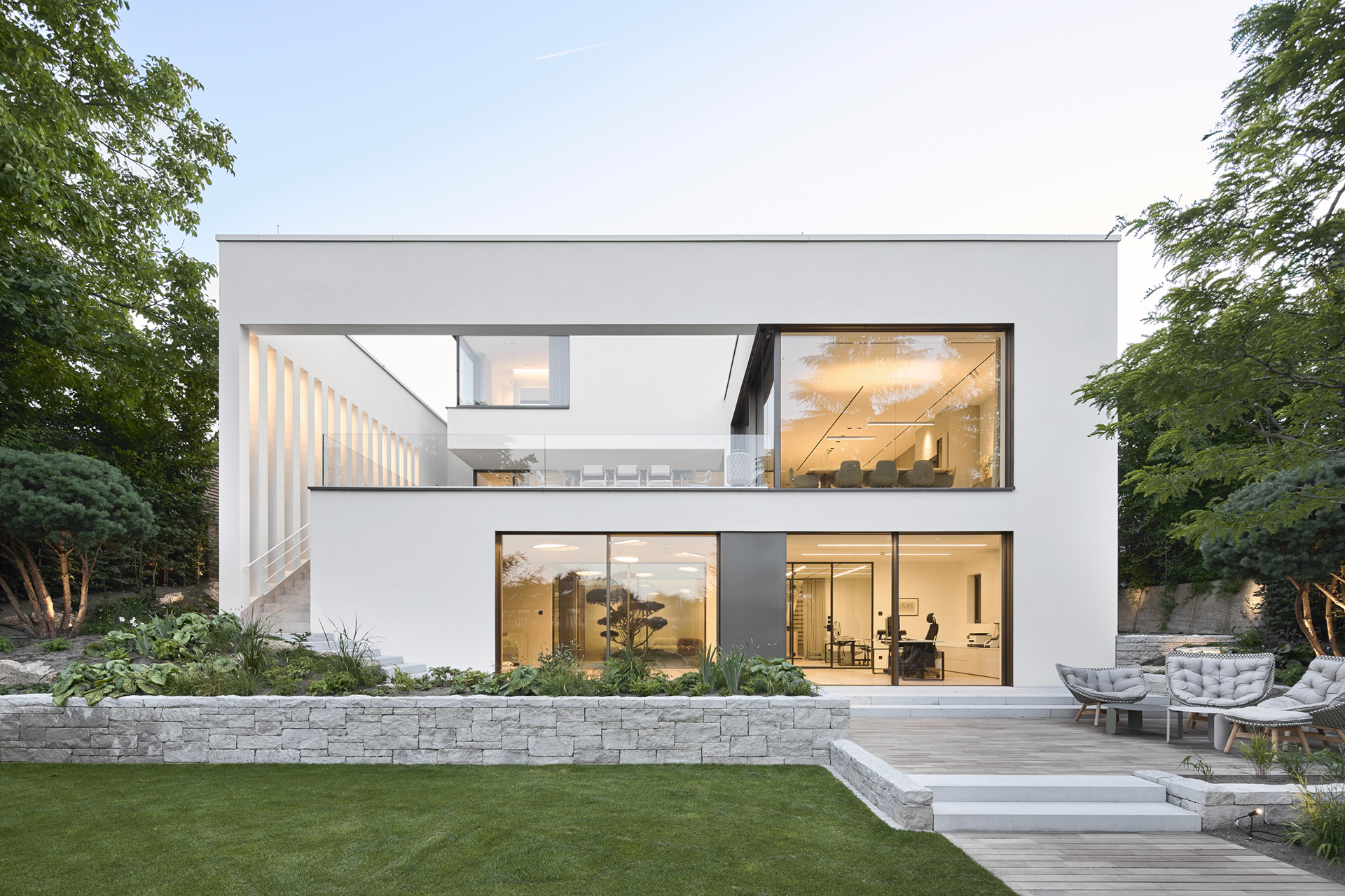 Diese Architekturfotografie zeigt ein modernes weises grosszügiges Einfamilienhaus mit grossen bodentiefen Fenstern im klassischen Stil von der Gartenseite aus in der Dämmerung.