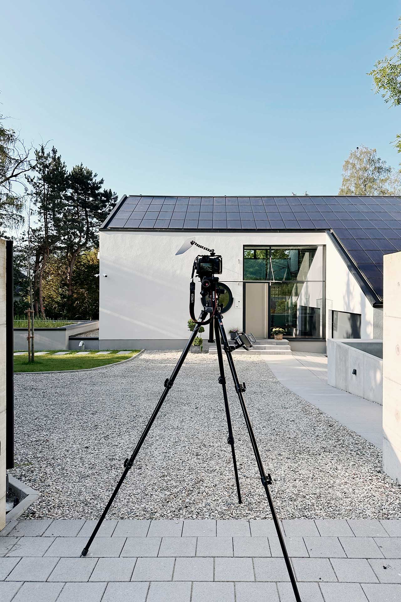 Architekturfotografie eines freistehenden Einfamilienhauses mit weissem Putz in Gevelsberg von der Eingangsseite mit Photovoltaik auf dem Dach bei Tageslicht und einer Kamera mit Stativ davor.