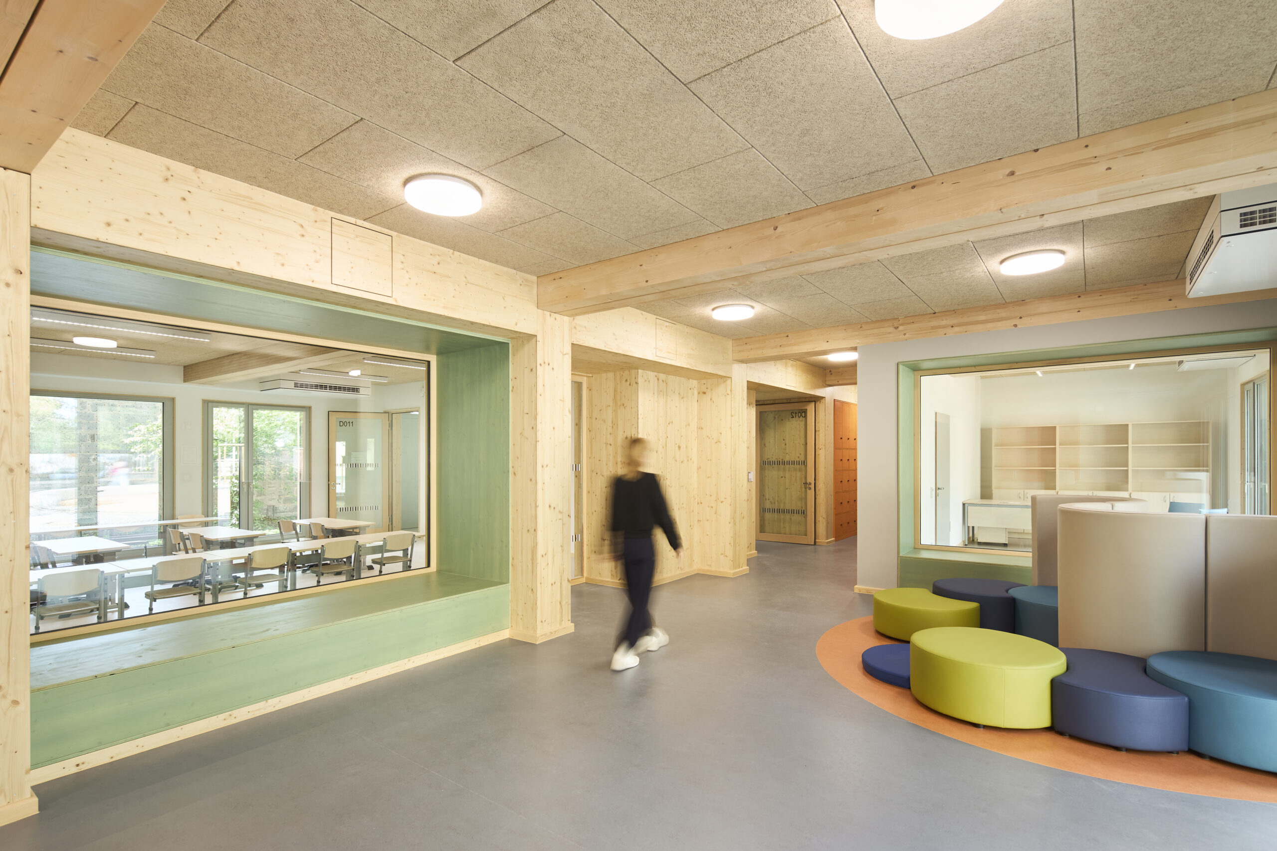 Diese Innenaufnahme zeigt den bunt gestalteten Flur einer Schule mit Holzausbauten, großem Fenster mit Durchblick in den Klassenraum und einer unscharfen Figur