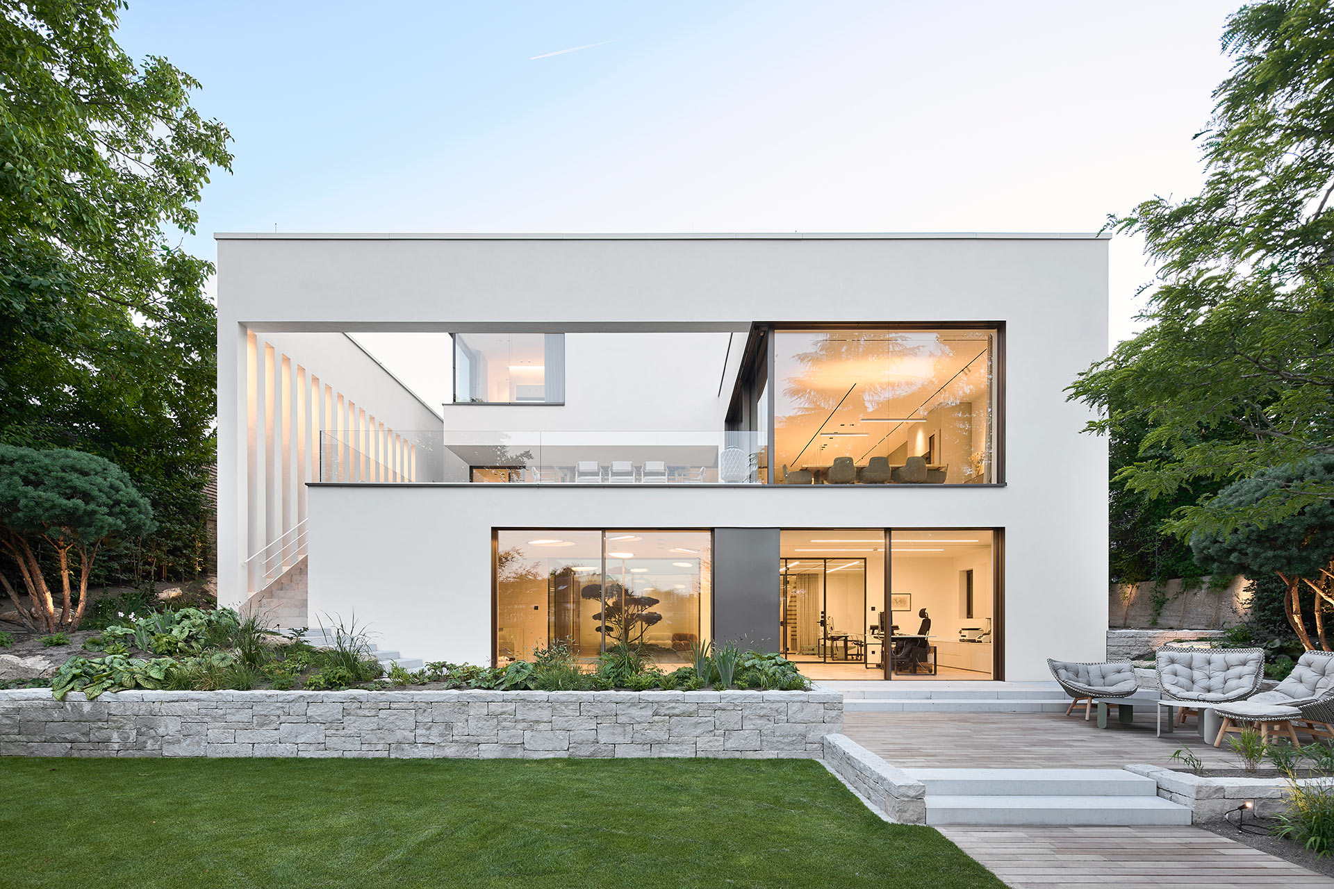 Diese Architekturfotografie zeigt ein modernes weises grosszügiges Einfamilienhaus mit grossen bodentiefen Fenstern im klassischen Stil von der Gartenseite aus in der Dämmerung.