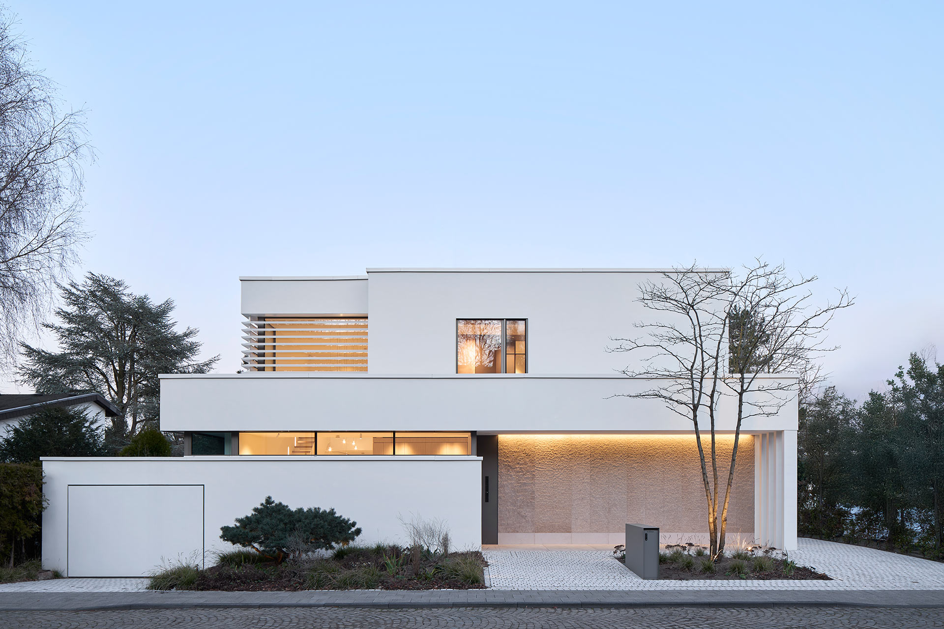 Diese Architekturfotografie zeigt ein modernes weises grosszügiges Einfamilienhaus mit zwei Etagen im klassischen Stil von der Straßenseite aus in der Dämmerung.