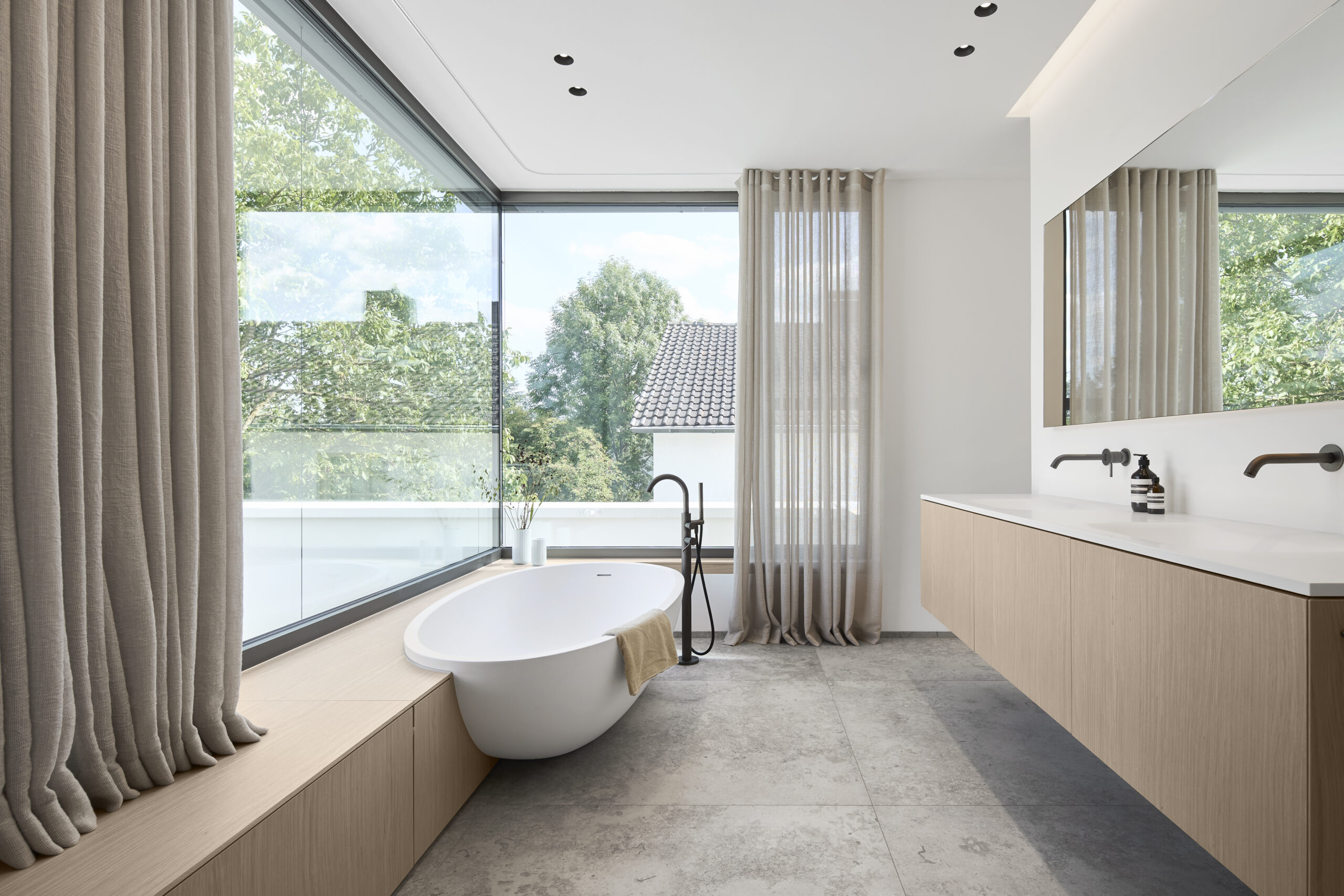 Diese Architekturfotografie zeigt das Badezimmer mit Verglasung über zwei Seiten, ovaler weisser Badewanne, die mittig in eine Holzverkleidung eingelassen ist. Der Waschtisch aus Holz und weisser Keramik erstreckt sich über beide Waschbecken.